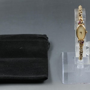 May include: A gold-tone analog wristwatch with an oval face and a twisted rope-style band. The watch features small, multi-colored gemstone accents above and below the dial. It is displayed on a clear stand next to a black velvet drawstring pouch.