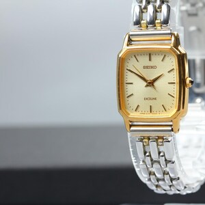 Puede incluir: Un reloj de pulsera Seiko Exceline con caja rectangular en tono dorado, esfera color champán, manecillas e índices dorados. El reloj cuenta con una correa de dos tonos compuesta por eslabones metálicos plateados y dorados alternos.