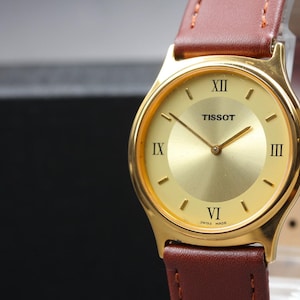 May include: A Tissot wristwatch with a round gold-tone dial, gold-tone hands, and black Roman numerals at the three, six, nine, and twelve o'clock positions. The watch features a polished gold-tone case and a brown leather strap with stitching.