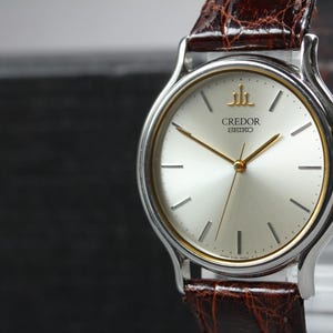 May include: A Credor Seiko wristwatch featuring a round silver dial with gold-tone hands and hour markers. The watch has a silver-tone bezel with a thin gold-tone inner ring and a brown textured leather strap with a glossy finish.