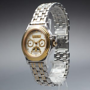 May include: A two-tone wristwatch with a silver and gold band and a white face. The watch face has gold accents, three sub-dials, and the text "STING Birthday Edition". The watch is displayed on a clear stand.