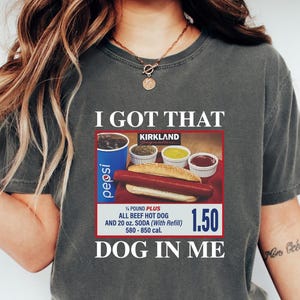 May include: Dark gray t-shirt with the text "I GOT THAT DOG IN ME." The shirt features a graphic of a hot dog, soda, and condiments. The shirt also has the text "KIRKLAND" and "1.50".