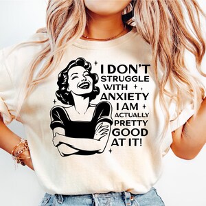 Könnte beinhalten: Cremefarbenes T-Shirt mit dem Text "I DON'T STRUGGLE WITH ANXIETY I AM ACTUALLY PRETTY GOOD AT IT!" und einer Schwarz-Weiß-Illustration einer lachenden Frau. Das Shirt wird mit hellblauen Jeans kombiniert.