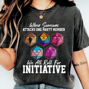 May include: Dark gray t-shirt with the text "When Someone Attacks One Party Member, We All Roll For Initiative." The shirt features colorful geometric dice with various symbols. The shirt is worn with blue jeans and white sneakers.