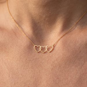 May include: A gold-colored necklace featuring three interconnected heart-shaped pendants. Each heart is engraved with a name in a cursive script: Emma, Leo, and Olivia. The pendant is suspended from a delicate, thin gold-colored chain.