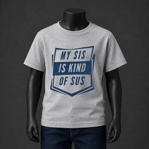 Puede incluir: Una camiseta de manga corta en color gris jaspeado con un diseño gráfico azul oscuro en el pecho. El gráfico tiene forma de escudo y contiene el texto MY SIS IS KIND OF SUS en una fuente sans-serif negrita con una textura desgastada.