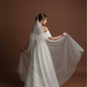 May include: A white wedding dress with an off-the-shoulder neckline and a flowing skirt. The bride is wearing a sheer veil that extends to the floor. The dress is made of a smooth, satin-like fabric. The background is a solid brown color.