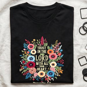 May include: A black short-sleeved t-shirt featuring a colourful floral wreath graphic surrounding the text "TRUST IN THE LORD WITH ALL YOUR HEART" and "Proverbs 3:5" printed in white and yellow lettering at the centre of the chest area.