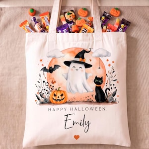 May include: A white canvas tote bag featuring a Halloween illustration of a ghost wearing a witch hat, a black cat, and a pumpkin against a large orange moon. The text reads Happy Halloween and the name Emily, with a small orange heart below.