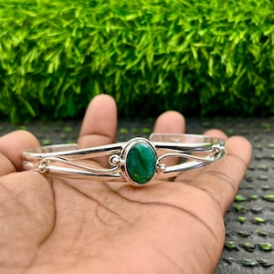 May include: A silver cuff bracelet with an oval, emerald-green stone set in the center. The bracelet has a delicate, open design with curved lines and small decorative accents. The bracelet is displayed on a hand against a green background.