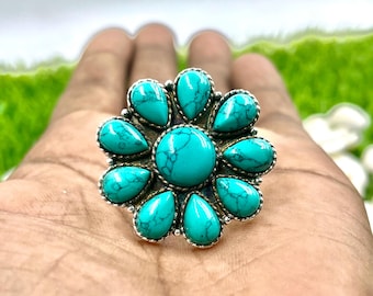Puede incluir: Un anillo plateado con un diseño floral, con una gran piedra turquesa redonda en el centro y once piedras turquesas en forma de lágrima. Las piedras del anillo están engastadas en biseles plateados, creando un contraste llamativo.