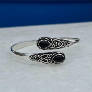 Op de afbeelding: Een zilverkleurige manchetarmband met twee zwarte, druppelvormige stenen. De armband heeft ingewikkelde details rond de stenen en langs de randen, met een decoratief patroon. Het ontwerp is open, waardoor het verstelbaar is.