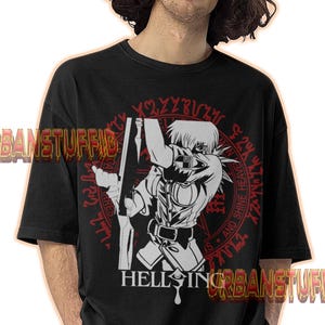 Puede incluir: Camiseta negra con un gráfico de anime blanco y rojo de un personaje con un rifle, con el texto "HELLSING" debajo. La camiseta también tiene el texto "URBANSTUFFID" en naranja. El diseño está sobre un patrón circular rojo.