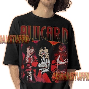 Puede incluir: Camiseta negra con la palabra "ALUCARD" en letras rojas y doradas. La camiseta muestra un gráfico de personajes de anime en rojo y negro. El texto "URBANSTUFFID" también es visible.