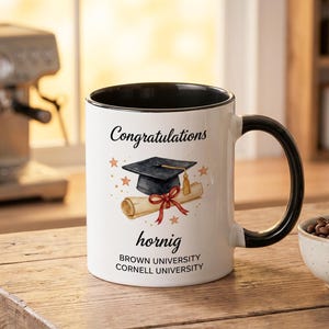 May include: White ceramic mug with a black handle and rim, featuring the text "Congratulations" and a graduation cap illustration. The mug also includes the name "hornig" and the university names "BROWN UNIVERSITY" and "CORNELL UNIVERSITY".