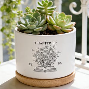 Puede incluir: Una maceta cilíndrica de cerámica blanca con base de madera que contiene suculentas verdes. La maceta presenta un gráfico de líneas negras de un libro abierto con un ramo de flores y mariposas, con el texto CHAPTER 30 y los años 19 y 96.