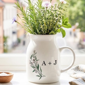 May include: A white ceramic pitcher with a handle, filled with fresh herbs and small blue and white flowers. The pitcher has a floral design and the letters "A + J" on the front. Other items on the windowsill include a glass jar, a small wooden bowl, and a salt shaker.