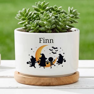 Op de afbeelding: Een witte keramische plantenpot met een bamboe schoteltje en een groene vetplant. De pot is bedrukt met de naam Finn boven een afbeelding van een gele maansikkel, zwarte vleermuizen en vier silhouetten van figuren in Halloween-kostuums.
