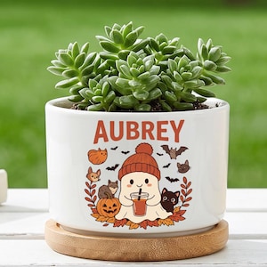 May include: A white ceramic planter with a wooden base containing a green succulent. The planter features the name AUBREY in orange letters above a graphic of a ghost wearing a beanie, holding a drink, surrounded by pumpkins, bats, and cats.