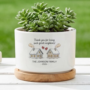 May include: A white ceramic cylindrical planter containing a green succulent plant, resting on a circular bamboo saucer. The planter features a graphic of two houses with smoke, red mittens, and the text: Thank you for being such great neighbours! THE JOHNSON FAMILY - 2026 -.