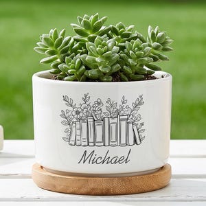 Peut inclure: Un pot de fleurs en céramique blanche avec une plante succulente, orné d'une illustration en noir et blanc de livres et de fleurs. Le nom "Michael" est imprimé. Il repose sur une soucoupe en bois.