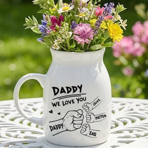 May include: A white ceramic jug with a handle, filled with a vibrant bouquet of colourful flowers. The jug has the text "DADDY WE LOVE YOU" and fist bump illustrations. The background is a blurred garden scene.