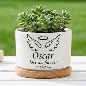 Op de afbeelding: Een witte keramische plantenpot met een groene vetplant, rustend op een ronde houten basis. De voorzijde toont een zwarte afbeelding van engelvleugels en een halo, met de tekst Oscar, love you forever en de jaartallen 2013-2026 eronder.