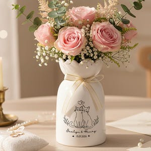 Op de afbeelding: Een witte keramische vaas met een boeket roze rozen, groen en gipskruid. De vaas heeft een kattenillustratie en de namen "Evelyn & Henry" met een datum. Een brandende kaars en een ringkussen zijn ook aanwezig.
