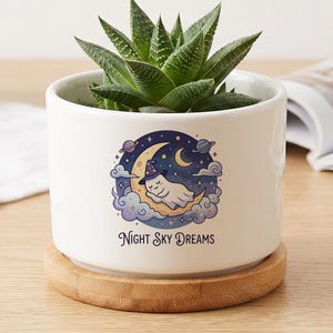 May include: A white ceramic cylindrical planter with a bamboo saucer base, containing a green succulent plant. The front features a graphic of a ghost wearing a wizard hat sleeping on a crescent moon, surrounded by stars, planets, and clouds, with the text "NIGHT SKY DREAMS" printed below.