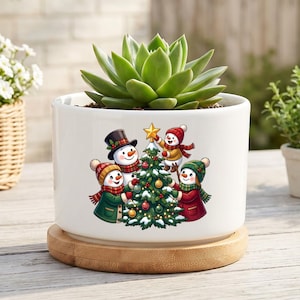 Peut inclure: Un pot de fleurs cylindrique en céramique blanche avec une base en bambou, contenant une plante succulente verte. L'avant du pot présente un graphique coloré de quatre bonhommes de neige décorant un sapin de Noël avec des boules et une étoile dorée.