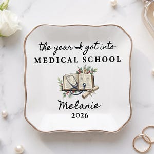 Peut inclure: Un vide-poche carré en céramique blanche avec des bords festonnés et un bord doré. Le centre présente le texte "the year I got into MEDICAL SCHOOL", une illustration d'un livre ouvert avec un stéthoscope et un diplôme, "Melanie" et "2026".