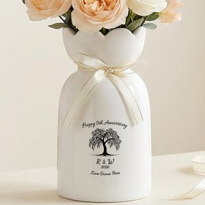 May include: A white ceramic vase with a bouquet of peach and white roses, and greenery. The vase has a unique shape and a ribbon tied around the neck. Text on the vase reads: "Happy 5th Anniversary 2020 R & W Love Grows Here."