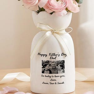 May include: A white vase with a scalloped top, tied with a ribbon, holds pink and white flowers. The vase has a photo and text that reads "Happy Father's Day Dad, So lucky to have you, Love, Liam, Thor & Sarah."