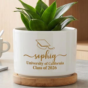 May include: A white ceramic cylindrical planter with a bamboo base containing a green plant. The planter features gold text that reads "sophig" above "University of California Class of 2026" and a gold line drawing of a graduation cap.