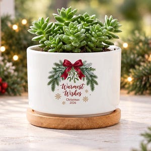 Könnte beinhalten: Ein weißer Keramikübertopf mit einer grünen Sukkulente auf einem runden Holzuntersetzer. Der Übertopf ist mit einer Grafik aus einer roten Schleife, Tannenzweigen und dem Text "Warmest Wishes Christmas 2026" in roter und goldener Schrift verziert.
