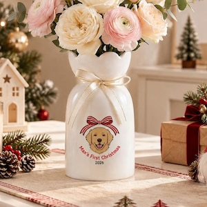 Puede incluir: Un jarrón de cerámica blanca con borde festoneado y una cinta de color crema atada alrededor del cuello. El frente presenta un gráfico de un golden retriever con un lazo a rayas rojas y blancas, con el texto Max's First Christmas 2026 debajo.