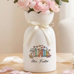 May include: A white ceramic vase filled with pink and white flowers, tied with a cream ribbon. The vase has a floral and book design with the text "Mrs. Foster". The vase is on a wooden table with scattered petals.