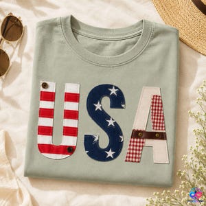 May include: A sage green crew neck t-shirt featuring the letters USA as large fabric appliqués. The U has red and white stripes with a brown button, the S is navy blue with white stars, and the A features red and white gingham with a brown leather strap.