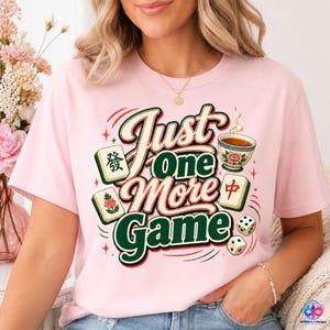 May include: A light pink t-shirt featuring a graphic design with the text "Just One More Game" in a retro font. The illustration includes mahjong tiles, a tea cup with a floral pattern, and two dice, all arranged in a colourful, decorative layout.