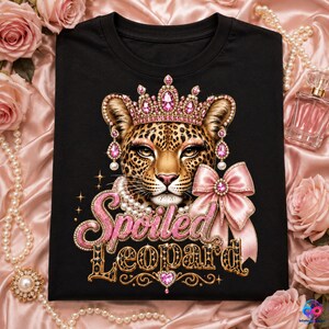 Op de afbeelding: Een zwart t-shirt met een afbeelding van een luipaard met een roze kroon vol juwelen en bijpassende oorbellen. De luipaard draagt een parelketting en een roze strik. Daaronder staat de tekst Spoiled Leopard in roze en gouden glitter.