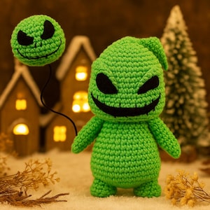 Może przedstawiać: Ręcznie robiona zielona lalka Oogie Boogie z szydełka z czarnymi oczami i wyszytym uśmiechem. Mniejsza, odłączona głowa unosi się nad nią, połączona czarnym sznurkiem. Lalka jest umieszczona na rozmytym tle z miniaturowymi domkami i choinką.