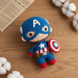 Puede incluir: Muñeco amigurumi de Capitán América hecho a mano. El muñeco es principalmente azul con una "A" blanca en la cabeza, una cara color beige y un escudo rojo, blanco y azul. El muñeco lleva un traje azul y blanco salpicado de estrellas con cinturón marrón y botas rojas.