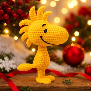 May include: A yellow crochet figure of Woodstock, the bird from Peanuts. The figure has a smiling face with black eyes and a beak. It is standing on a wooden surface with Christmas decorations in the background.