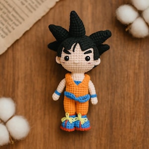 Puede incluir: Muñeca de ganchillo hecha a mano de un personaje con pelo negro puntiagudo, vistiendo un atuendo naranja con detalles azules. La muñeca tiene un tono de piel neutro y está colocada sobre un fondo de madera. Los zapatos de la muñeca son azules y rojos con detalles amarillos.