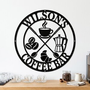 Puede incluir: Un letrero de pared circular de metal negro con el texto WILSON'S COFFEE BAR. El diseño incluye una taza de café con vapor, una cafetera moka, granos de café y un cruasán, enmarcados por dos cucharas cruzadas dentro del borde circular.