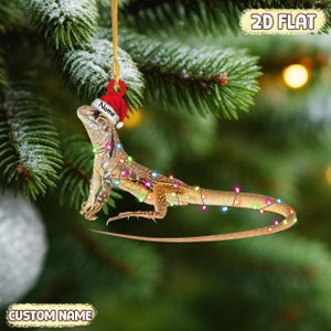 May include: A lizard-shaped Christmas ornament with a red Santa hat and string lights. The ornament is brown and tan with a long tail. The text "Name" is on the hat. The text "2D FLAT" and "CUSTOM NAME" are also visible.