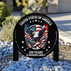 Puede incluir: Estaca de jardín de metal negro con un diseño patriótico. Muestra un águila calva, la bandera estadounidense y el texto "UNITED STATES OF AMERICA" y "250 YEARS OF DEFENDING LIBERTY". La estaca está colocada en un lecho de pequeñas rocas.