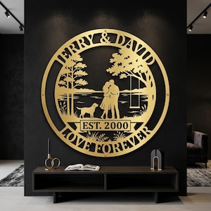 Puede incluir: Una pieza de arte mural de metal dorado con los nombres "JERRY & DAVID" en un diseño circular. El arte presenta una pareja, un perro, árboles y el texto "EST. 2000 LOVE FOREVER". Está montado en una pared negra sobre una consola de madera oscura.