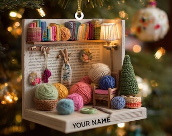 May include: A miniature Christmas ornament featuring a cozy scene with yarn, books, and a small lamp. The ornament includes a miniature chair, a small green tree, and the text "YOUR NAME". The ornament is suspended by a gold string.