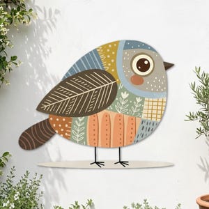 May include: A decorative wall art piece shaped like a plump bird, featuring a patchwork design of muted blue, mustard yellow, brown, and peach sections. The bird has a large circular eye, a dark brown wing with leaf patterns, and thin black legs.